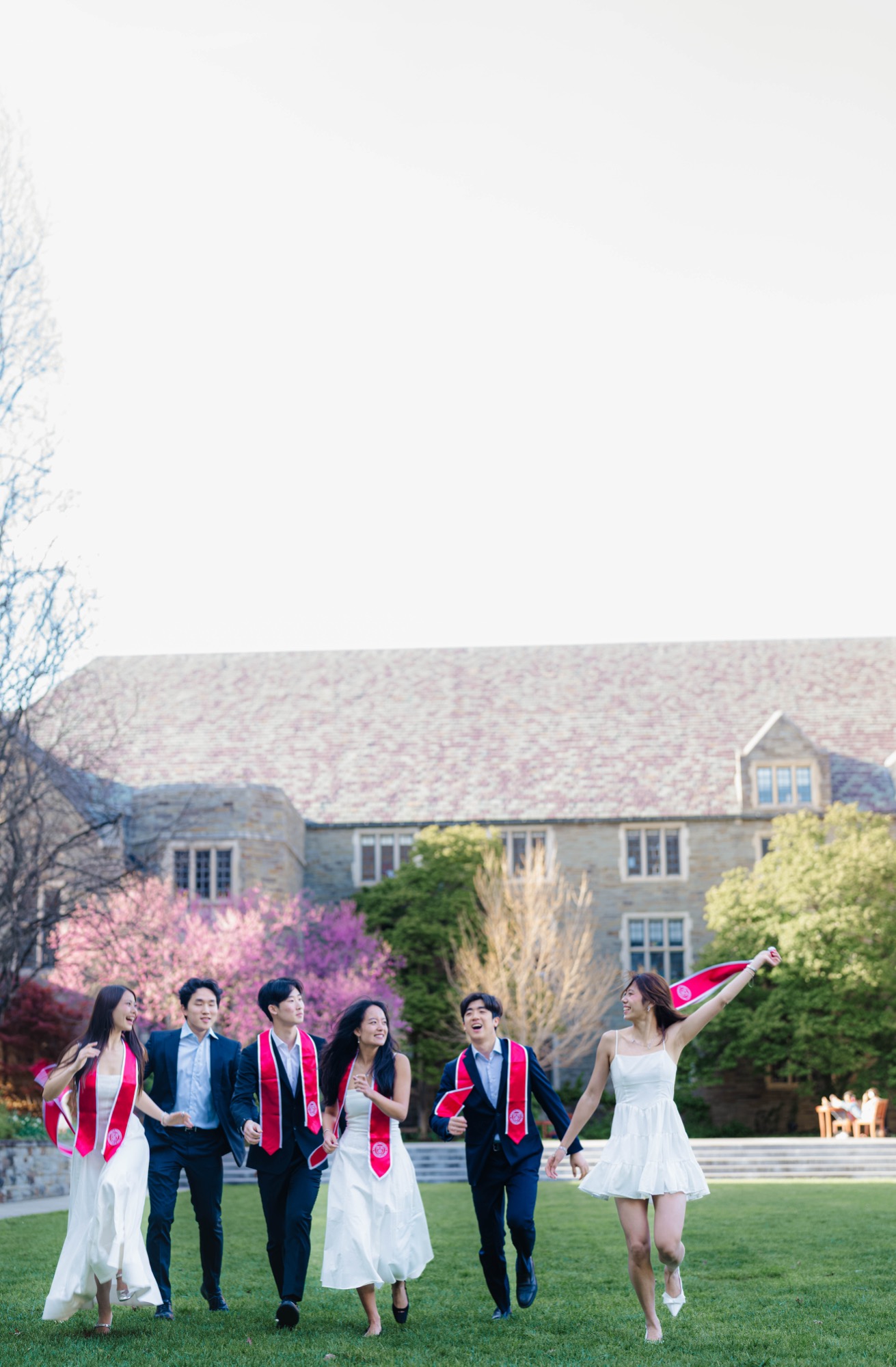 Six graduating members walking together across the lawn, redbuds in bloom and a stone hall behind them